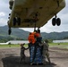 Military and civilian personnel are attaching a cargo net-wrapped pallet of supplies to the underside of a large military helicopter, likely a CH-47 Chinook, on a tarmac. The operation appears to be a sling-load or external lift preparation for humanitarian aid delivery. The lush green mountainous terrain in the background suggests a tropical location.