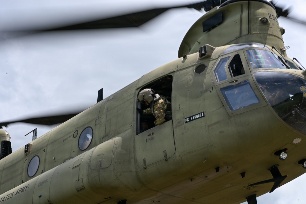 A U.S. Army CH-47 Chinook helicopter is captured in flight at close range, showing the forward fuselage section in olive drab livery. A crew member wearing a flight helmet and tactical gear is visible leaning out of the forward crew door window. The name placard 'FE VASQUEZ' is clearly visible on the fuselage, indicating the flight engineer assigned to this aircraft.