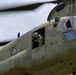 A U.S. Army CH-47 Chinook helicopter is captured in flight at close range, showing the forward fuselage section in olive drab livery. A crew member wearing a flight helmet and tactical gear is visible leaning out of the forward crew door window. The name placard 'FE VASQUEZ' is clearly visible on the fuselage, indicating the flight engineer assigned to this aircraft.