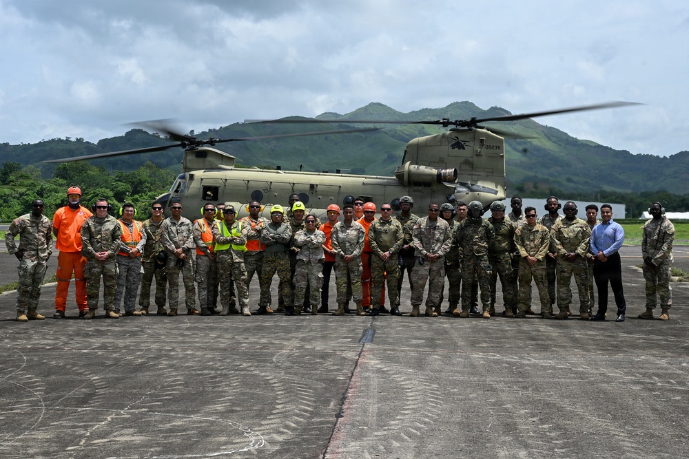 A large group of approximately 25-30 military personnel and civilian workers pose for a group photo in front of a CH-47 Chinook helicopter on a tarmac or landing pad. The setting appears to be a tropical or subtropical location, evidenced by the lush green mountains in the background. The group includes both uniformed military members in camouflage and civilian workers wearing high-visibility safety gear.