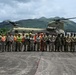 A large group of approximately 25-30 military personnel and civilian workers pose for a group photo in front of a CH-47 Chinook helicopter on a tarmac or landing pad. The setting appears to be a tropical or subtropical location, evidenced by the lush green mountains in the background. The group includes both uniformed military members in camouflage and civilian workers wearing high-visibility safety gear.