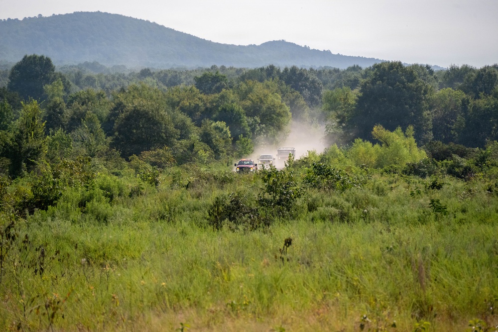 A convoy of vehicles is traveling along a dusty dirt road through a rural, heavily vegetated landscape. The vehicles are kicking up a visible dust cloud as they move through the terrain, surrounded by dense green shrubs and trees. A forested hillside and a low mountain ridge are visible in the hazy background.