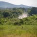 A convoy of vehicles is traveling along a dusty dirt road through a rural, heavily vegetated landscape. The vehicles are kicking up a visible dust cloud as they move through the terrain, surrounded by dense green shrubs and trees. A forested hillside and a low mountain ridge are visible in the hazy background.