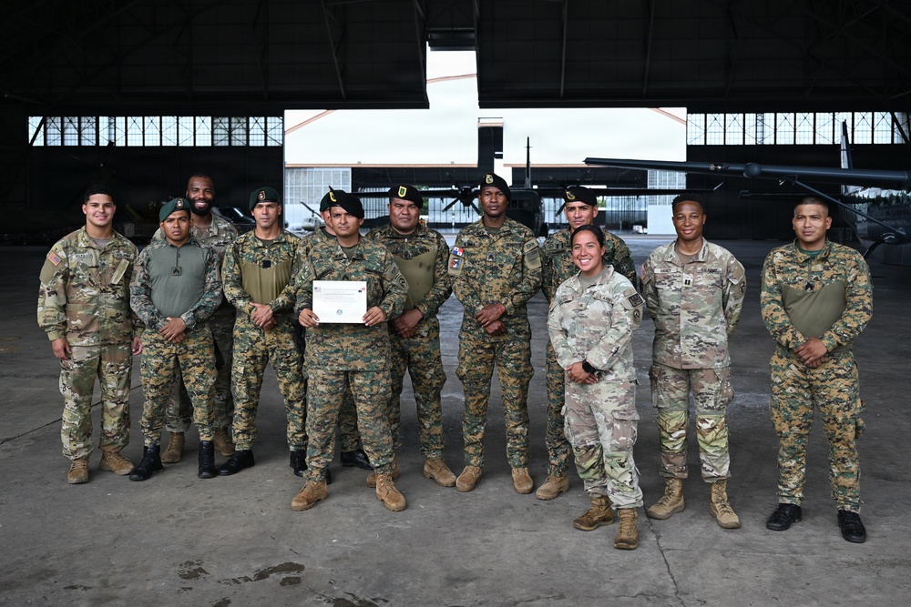 A group of approximately 12 military personnel pose for a formal group photograph inside an aircraft hangar. The group includes both U.S. Army soldiers in MultiCam/OCP uniforms and foreign military personnel in woodland digital camouflage uniforms with black berets. One individual in the center is holding what appears to be a certificate or document, suggesting a graduation, training completion, or award ceremony.