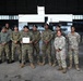 A group of approximately 12 military personnel pose for a formal group photograph inside an aircraft hangar. The group includes both U.S. Army soldiers in MultiCam/OCP uniforms and foreign military personnel in woodland digital camouflage uniforms with black berets. One individual in the center is holding what appears to be a certificate or document, suggesting a graduation, training completion, or award ceremony.