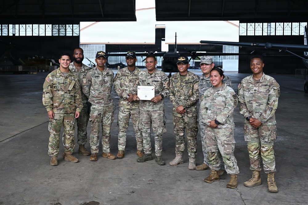 A group of nine military personnel in camouflage uniforms pose for a formal group photo inside a large aircraft hangar. One individual in the center is holding what appears to be a certificate or award document, suggesting a recognition or graduation ceremony. The setting includes military helicopters visible in the background of the hangar.