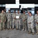 A group of nine military personnel in camouflage uniforms pose for a formal group photo inside a large aircraft hangar. One individual in the center is holding what appears to be a certificate or award document, suggesting a recognition or graduation ceremony. The setting includes military helicopters visible in the background of the hangar.