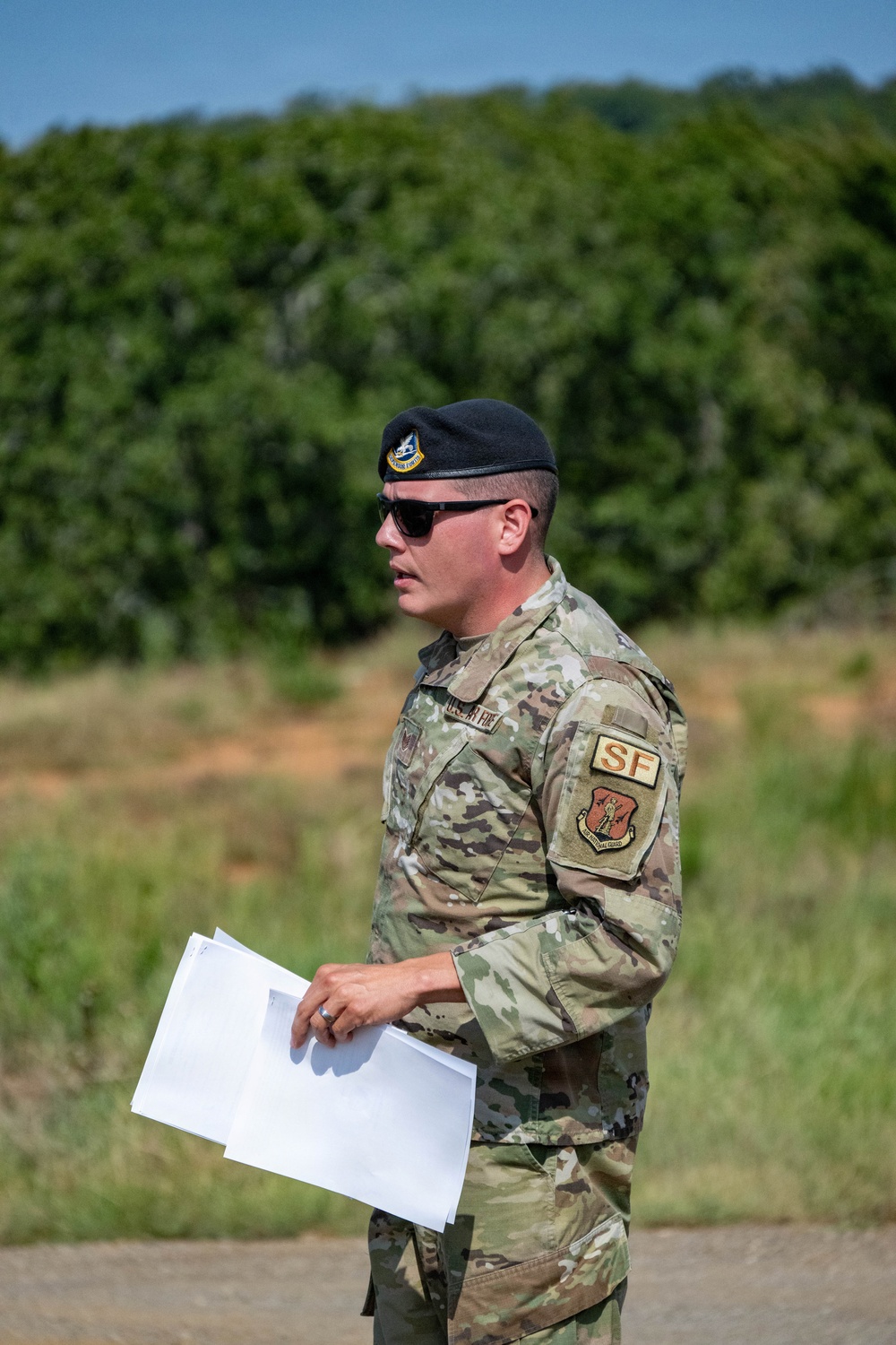 A U.S. Air Force Security Forces (SF) member stands outdoors in a field or training area, wearing OCP (Operational Camouflage Pattern) uniform and a black beret. He is holding several sheets of white paper and appears to be speaking or briefing. Patches on his sleeve indicate Security Forces (SF) designation and a unit shield patch consistent with Air National Guard.