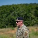 A U.S. Air Force Security Forces (SF) member stands outdoors in a field or training area, wearing OCP (Operational Camouflage Pattern) uniform and a black beret. He is holding several sheets of white paper and appears to be speaking or briefing. Patches on his sleeve indicate Security Forces (SF) designation and a unit shield patch consistent with Air National Guard.
