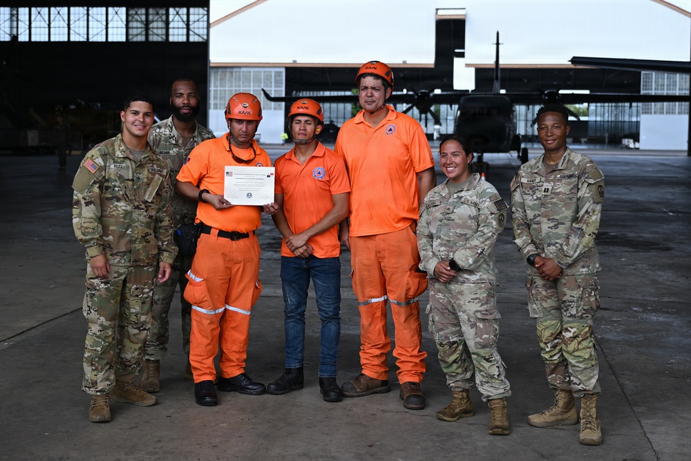 A group of seven individuals pose for a formal photo in front of a military hangar with a helicopter visible in the background. The group consists of four U.S. Army soldiers in OCP (Operational Camouflage Pattern) uniforms and three civilians wearing orange emergency response uniforms and helmets. One of the civilians is holding a certificate, suggesting a completion or recognition ceremony.