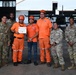 A group of seven individuals pose for a formal photo in front of a military hangar with a helicopter visible in the background. The group consists of four U.S. Army soldiers in OCP (Operational Camouflage Pattern) uniforms and three civilians wearing orange emergency response uniforms and helmets. One of the civilians is holding a certificate, suggesting a completion or recognition ceremony.