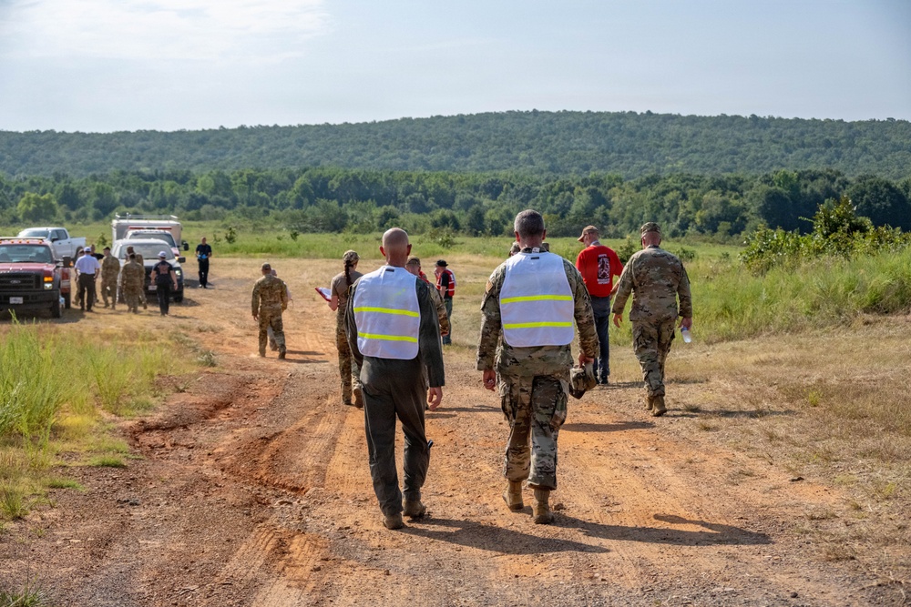 A group of military personnel and civilians are walking along a dirt road in a rural, open field setting with forested hills in the background. Two individuals in the foreground are wearing white reflective safety vests over military camouflage uniforms, suggesting they may be observers or evaluators during a training exercise or field operation. Several vehicles, including a white truck, are visible in the background where additional personnel are gathered.
