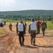 A group of military personnel and civilians are walking along a dirt road in a rural, open field setting with forested hills in the background. Two individuals in the foreground are wearing white reflective safety vests over military camouflage uniforms, suggesting they may be observers or evaluators during a training exercise or field operation. Several vehicles, including a white truck, are visible in the background where additional personnel are gathered.