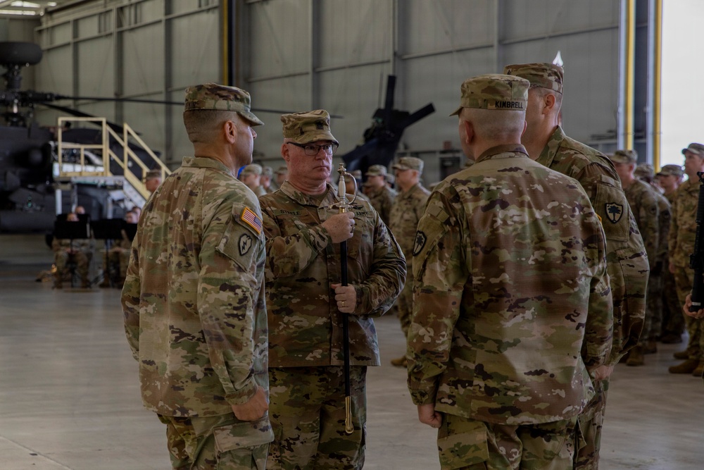 A military ceremony is taking place inside a large aircraft hangar, with U.S. Army soldiers in OCP (Operational Camouflage Pattern) uniforms gathered in formation. A central soldier, identified by nametape as 'STEELE,' is holding a ceremonial saber, suggesting this is a change of command or similar formal military ceremony. Apache helicopters are visible in the background of the hangar.