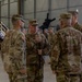 A military ceremony is taking place inside a large aircraft hangar, with U.S. Army soldiers in OCP (Operational Camouflage Pattern) uniforms gathered in formation. A central soldier, identified by nametape as 'STEELE,' is holding a ceremonial saber, suggesting this is a change of command or similar formal military ceremony. Apache helicopters are visible in the background of the hangar.