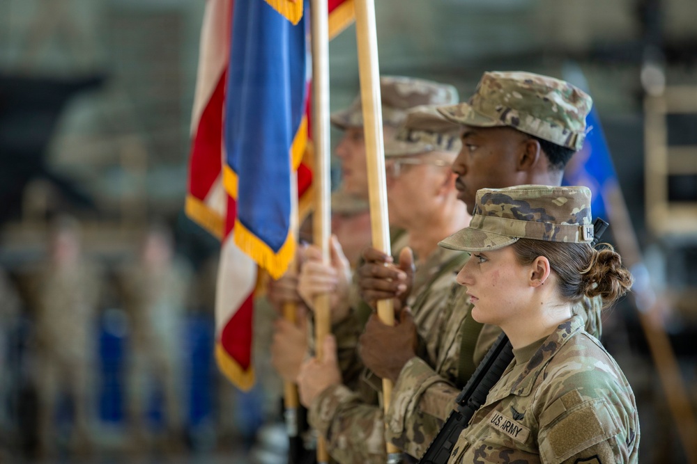 A U.S. Army color guard is participating in a formal military ceremony, holding flags with gold fringe in what appears to be a hangar or large indoor facility. Multiple soldiers in OCP (Operational Camouflage Pattern) uniforms are standing at attention in formation. The foreground soldier is clearly identified as U.S. Army personnel with a name tape partially visible.