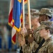 A U.S. Army color guard is participating in a formal military ceremony, holding flags with gold fringe in what appears to be a hangar or large indoor facility. Multiple soldiers in OCP (Operational Camouflage Pattern) uniforms are standing at attention in formation. The foreground soldier is clearly identified as U.S. Army personnel with a name tape partially visible.