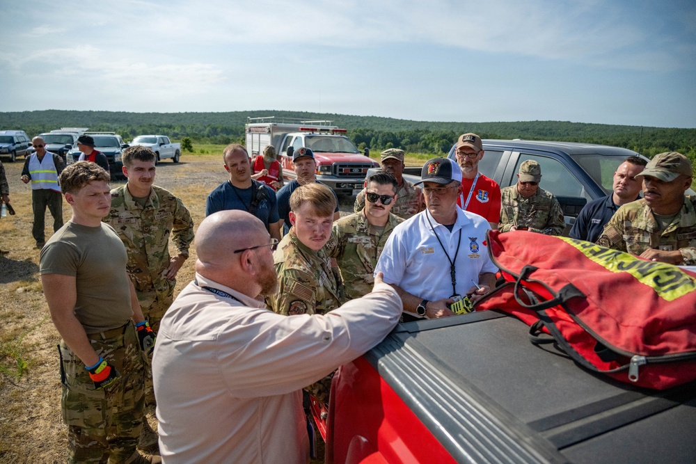A group of military personnel in camouflage uniforms and civilian instructors in white shirts gather around the bed of a red pickup truck in an outdoor field setting. A bald instructor with a red beard appears to be conducting a briefing or equipment demonstration, with emergency/rescue gear visible on the truck bed. The setting suggests a training exercise or field operation, with a fire truck and other vehicles visible in the background.