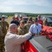 A group of military personnel in camouflage uniforms and civilian instructors in white shirts gather around the bed of a red pickup truck in an outdoor field setting. A bald instructor with a red beard appears to be conducting a briefing or equipment demonstration, with emergency/rescue gear visible on the truck bed. The setting suggests a training exercise or field operation, with a fire truck and other vehicles visible in the background.
