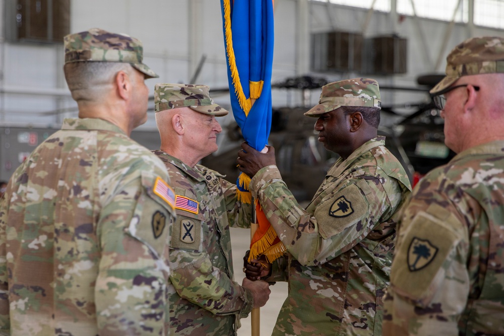 A military change of command ceremony is taking place inside what appears to be an aircraft hangar. Two central figures are exchanging a blue guidon/unit flag with gold fringe, which is the traditional symbolic act of transferring command authority. At least four U.S. Army soldiers in OCP (Operational Camouflage Pattern) uniforms are present, with military helicopters visible in the background.