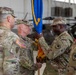 A military change of command ceremony is taking place inside what appears to be an aircraft hangar. Two central figures are exchanging a blue guidon/unit flag with gold fringe, which is the traditional symbolic act of transferring command authority. At least four U.S. Army soldiers in OCP (Operational Camouflage Pattern) uniforms are present, with military helicopters visible in the background.