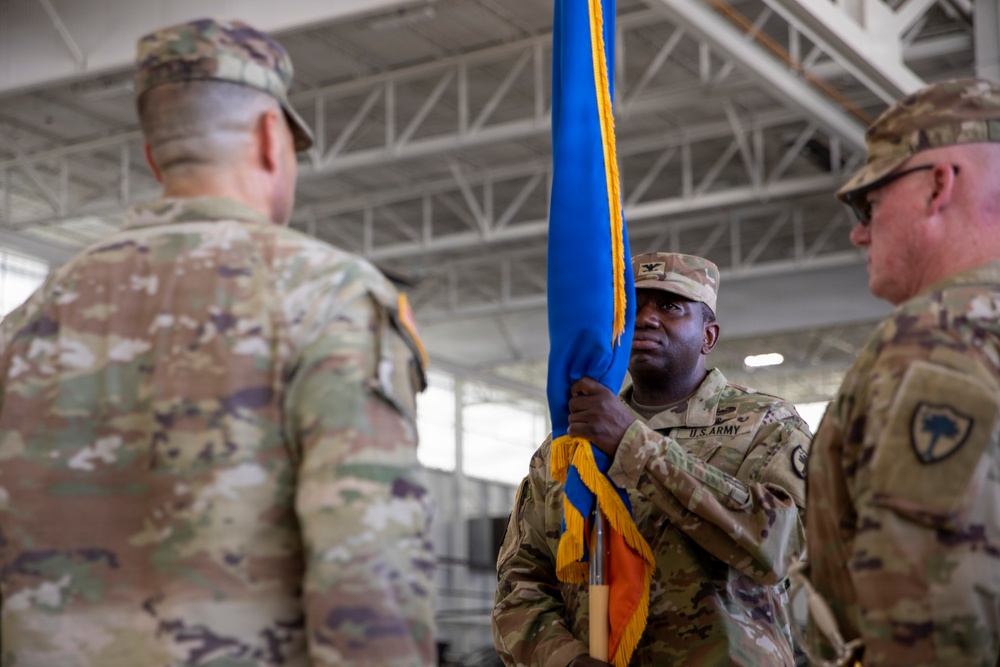 A U.S. Army change of command or activation ceremony is taking place inside what appears to be a military hangar or large indoor facility. A soldier in the center, wearing a U.S. Army uniform with a colonel's eagle insignia, is holding a blue and gold guidon or unit colors. Two other soldiers flank him, one facing away from the camera and one visible in profile on the right.