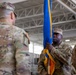 A U.S. Army change of command or activation ceremony is taking place inside what appears to be a military hangar or large indoor facility. A soldier in the center, wearing a U.S. Army uniform with a colonel's eagle insignia, is holding a blue and gold guidon or unit colors. Two other soldiers flank him, one facing away from the camera and one visible in profile on the right.