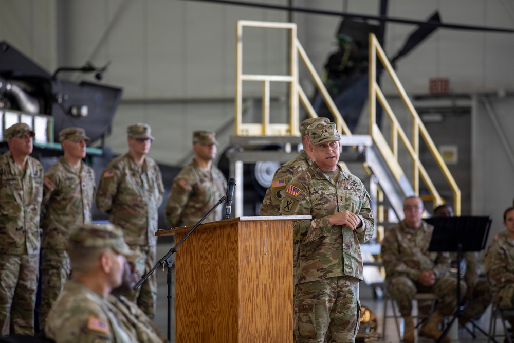 A U.S. Army officer is delivering a speech at a podium during what appears to be a formal military ceremony held inside an aircraft hangar. Multiple soldiers in Army Combat Uniforms (ACUs) in Operational Camouflage Pattern (OCP) are standing in formation in the background, while others are seated to the right. The setting suggests a change of command, unit activation, or similar official military ceremony.