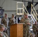 A U.S. Army officer is delivering a speech at a podium during what appears to be a formal military ceremony held inside an aircraft hangar. Multiple soldiers in Army Combat Uniforms (ACUs) in Operational Camouflage Pattern (OCP) are standing in formation in the background, while others are seated to the right. The setting suggests a change of command, unit activation, or similar official military ceremony.