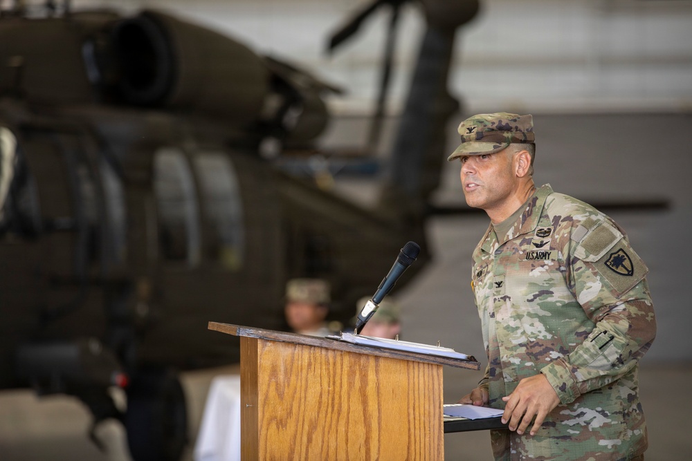 A U.S. Army officer in OCP (Operational Camouflage Pattern) uniform is speaking at a wooden podium with a microphone, likely during a military ceremony or official event. The setting appears to be inside a military hangar, with a UH-60 Black Hawk helicopter visible in the background. A second uniformed individual is partially visible seated behind the speaker.