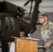 A U.S. Army officer in OCP (Operational Camouflage Pattern) uniform is speaking at a wooden podium with a microphone, likely during a military ceremony or official event. The setting appears to be inside a military hangar, with a UH-60 Black Hawk helicopter visible in the background. A second uniformed individual is partially visible seated behind the speaker.