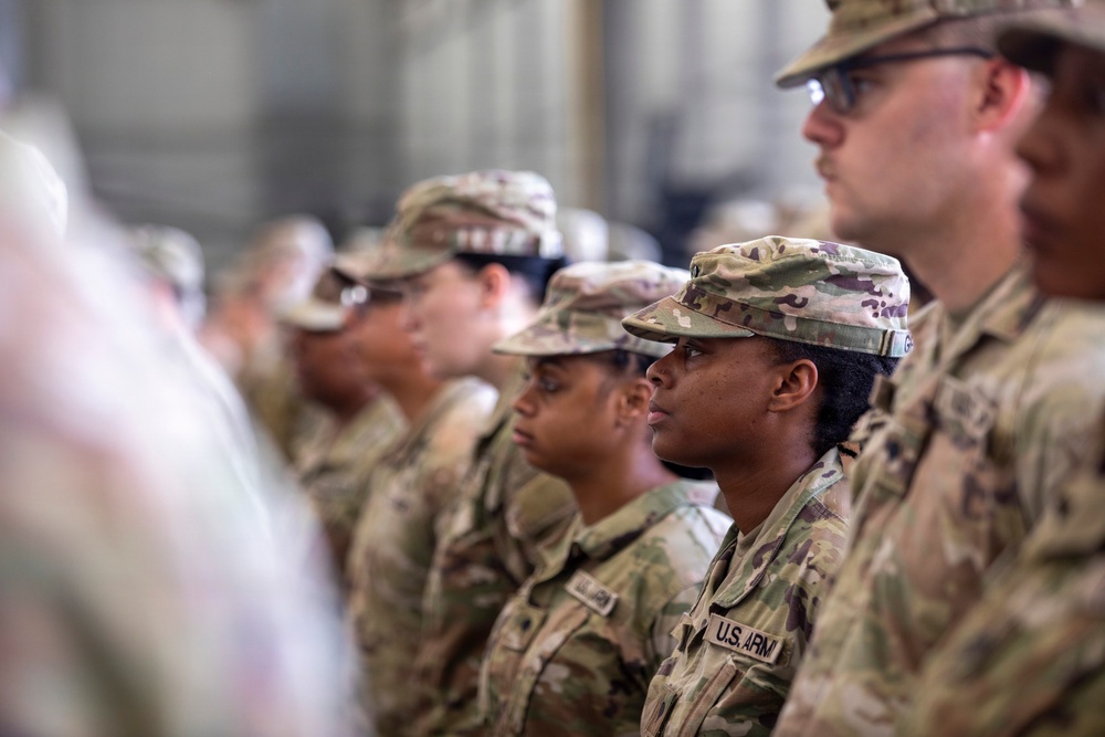 A group of U.S. Army soldiers standing in formation, wearing Operational Camouflage Pattern (OCP) uniforms. The soldiers appear to be at attention during a formal military ceremony or briefing held indoors. Both male and female soldiers of diverse backgrounds are visible in the formation.
