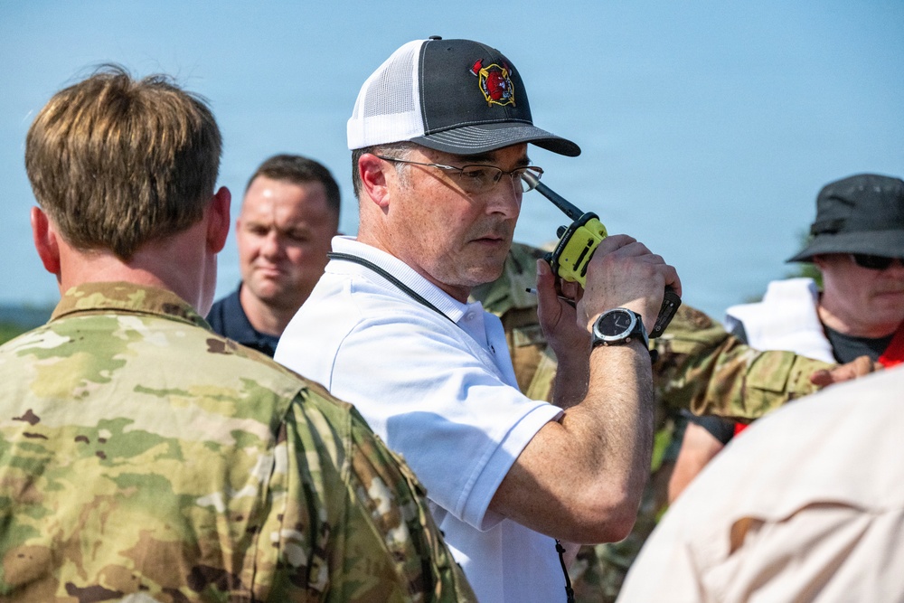 A group of individuals, including military personnel in camouflage uniforms and civilians, are gathered outdoors in what appears to be an emergency management or coordination setting. The central figure, wearing a white polo shirt and a fire department baseball cap, is holding a yellow-green handheld radio to his ear. The scene suggests a multi-agency coordination event, exercise, or active incident response.