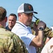 A group of individuals, including military personnel in camouflage uniforms and civilians, are gathered outdoors in what appears to be an emergency management or coordination setting. The central figure, wearing a white polo shirt and a fire department baseball cap, is holding a yellow-green handheld radio to his ear. The scene suggests a multi-agency coordination event, exercise, or active incident response.