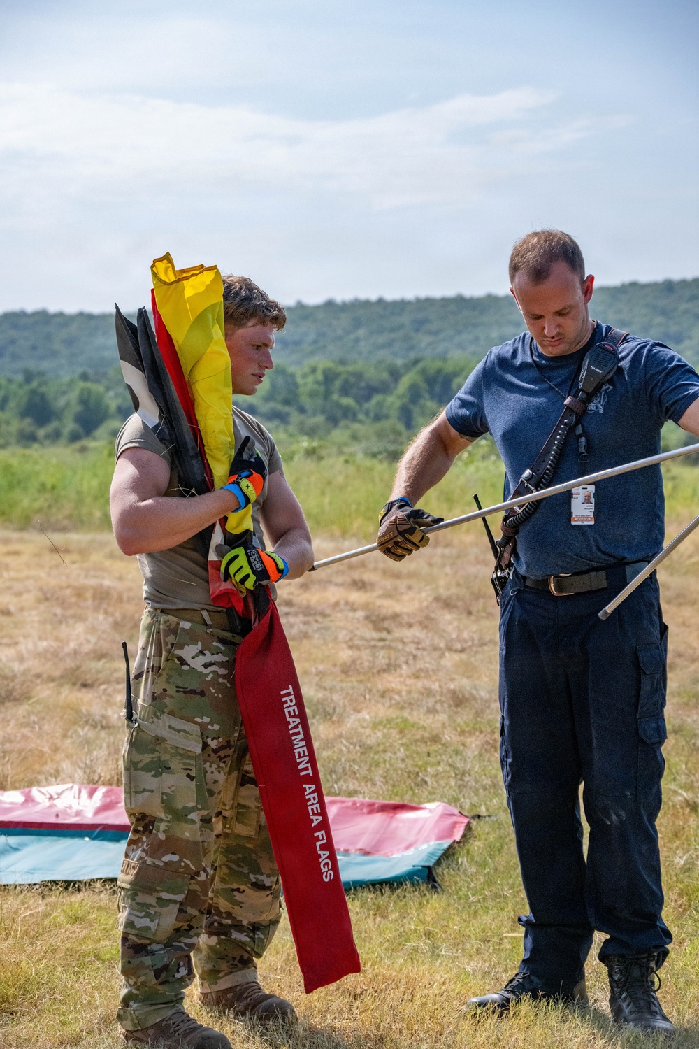 Two individuals are working together in an open field, handling equipment including colored flags and metal poles. One person is wearing military camouflage uniform and tactical gloves while carrying multiple flags, including a red one labeled 'TREATMENT AREA FLAGS' and a yellow flag. The other individual is in civilian/emergency services attire with a blue shirt, dark pants, tactical gloves, and a visible ID badge.