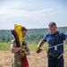 Two individuals are working together in an open field, handling equipment including colored flags and metal poles. One person is wearing military camouflage uniform and tactical gloves while carrying multiple flags, including a red one labeled 'TREATMENT AREA FLAGS' and a yellow flag. The other individual is in civilian/emergency services attire with a blue shirt, dark pants, tactical gloves, and a visible ID badge.