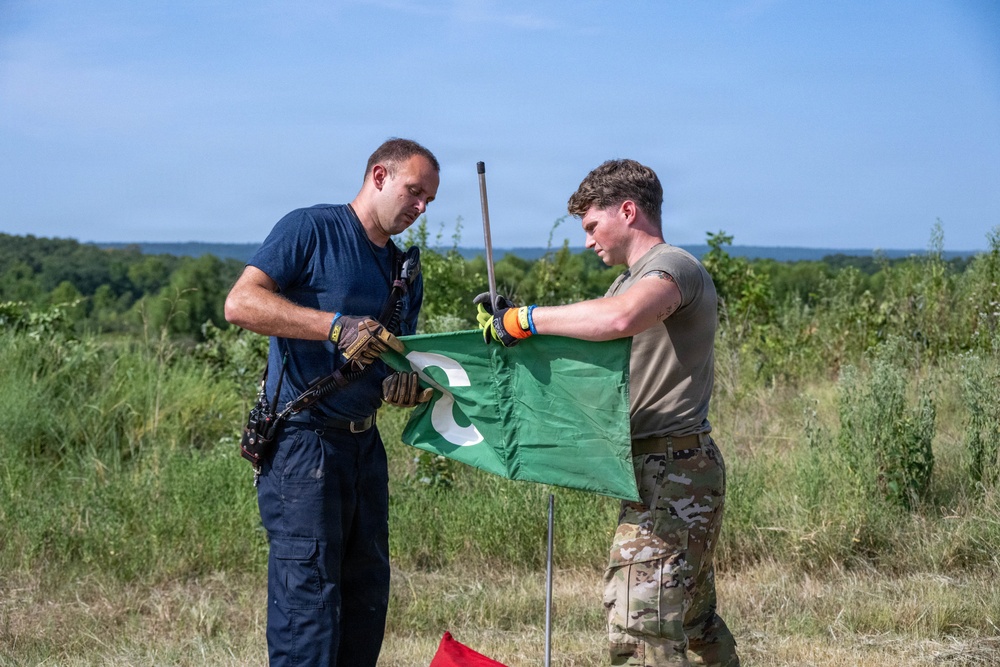 Two individuals, one in dark navy tactical clothing and one in military multicam uniform, are handling a green flag mounted on a metal pole in an outdoor field setting. Both are wearing tactical gloves and appear to be either setting up or taking down the flag. The setting appears to be a training range or exercise area based on the terrain and attire.