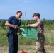 Two individuals, one in dark navy tactical clothing and one in military multicam uniform, are handling a green flag mounted on a metal pole in an outdoor field setting. Both are wearing tactical gloves and appear to be either setting up or taking down the flag. The setting appears to be a training range or exercise area based on the terrain and attire.