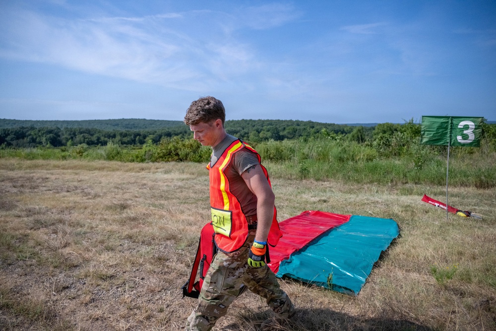 A young male in military camouflage pants and a high-visibility orange safety vest is walking across an open grassy field. He appears to be participating in a military training exercise or competition, evidenced by the numbered green flag marker (station 3) and colored mat visible in the background. The setting is an outdoor training area with treeline in the distance under clear summer skies.
