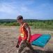 A young male in military camouflage pants and a high-visibility orange safety vest is walking across an open grassy field. He appears to be participating in a military training exercise or competition, evidenced by the numbered green flag marker (station 3) and colored mat visible in the background. The setting is an outdoor training area with treeline in the distance under clear summer skies.
