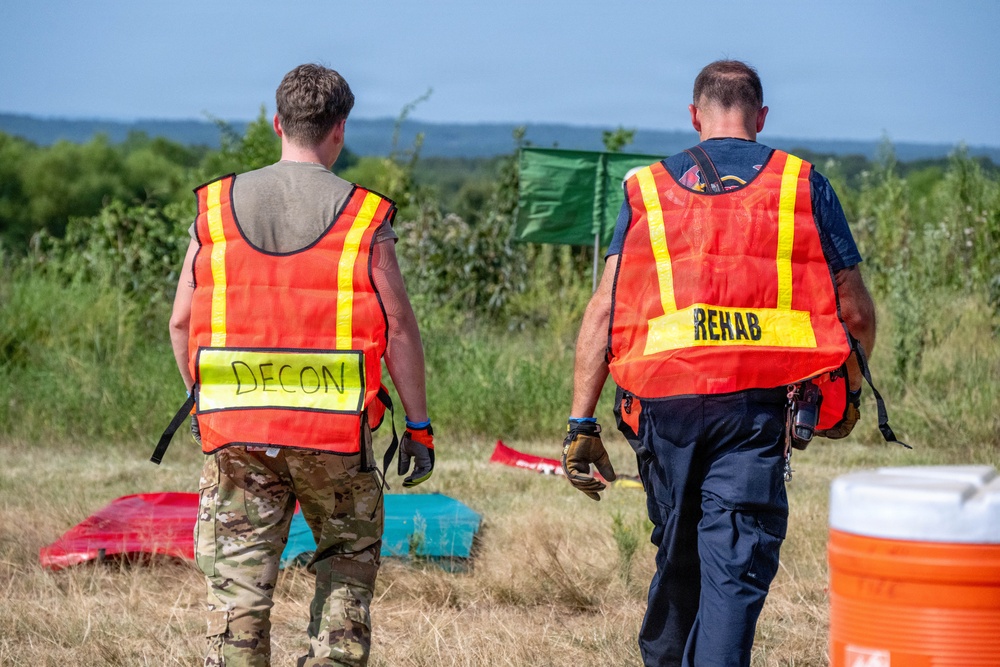 Two personnel wearing high-visibility orange safety vests are walking away from the camera across a grassy field. The vest on the left is labeled 'DECON' (decontamination) and the vest on the right is labeled 'REHAB' (rehabilitation), indicating this is an emergency response exercise or incident. The setting appears to be an outdoor training or operational area with equipment visible on the ground.