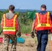 Two personnel wearing high-visibility orange safety vests are walking away from the camera across a grassy field. The vest on the left is labeled 'DECON' (decontamination) and the vest on the right is labeled 'REHAB' (rehabilitation), indicating this is an emergency response exercise or incident. The setting appears to be an outdoor training or operational area with equipment visible on the ground.