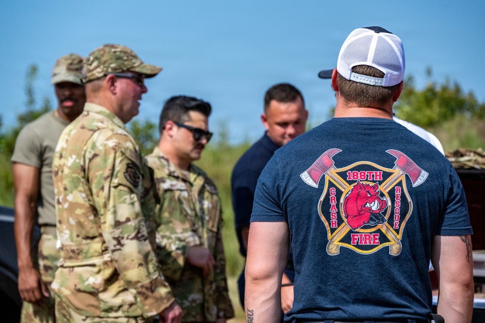 A group of individuals are gathered outdoors, including military personnel in MultiCam/OCP camouflage uniforms and civilians. The foreground figure wears a navy blue t-shirt displaying '188th Crash Fire Rescue' insignia with crossed axes and a boar mascot. The setting appears to be a field or outdoor training/operational area under clear blue skies.