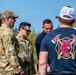 A group of individuals are gathered outdoors, including military personnel in MultiCam/OCP camouflage uniforms and civilians. The foreground figure wears a navy blue t-shirt displaying '188th Crash Fire Rescue' insignia with crossed axes and a boar mascot. The setting appears to be a field or outdoor training/operational area under clear blue skies.