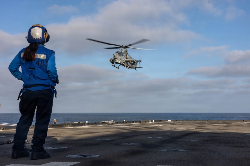 A naval flight deck crew member in a blue jersey and helmet stands on the flight deck of a warship observing a military helicopter in a hover or landing approach. The helicopter appears to be a Bell AH-1Z Viper attack helicopter, identifiable by its tandem cockpit and weapons systems. The scene takes place at sea under partly cloudy skies.