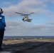 A naval flight deck crew member in a blue jersey and helmet stands on the flight deck of a warship observing a military helicopter in a hover or landing approach. The helicopter appears to be a Bell AH-1Z Viper attack helicopter, identifiable by its tandem cockpit and weapons systems. The scene takes place at sea under partly cloudy skies.