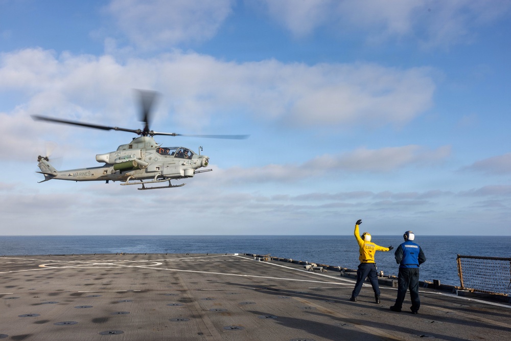 A U.S. Marines AH-1Z Viper attack helicopter is hovering just above the flight deck of a naval vessel, likely an amphibious assault ship, during flight operations at sea. Two deck crew members — one in a yellow jersey and one in a blue jersey — are directing the aircraft, with the yellow-jersey handler raising one arm in a signaling gesture. The scene takes place under partly cloudy skies over open ocean.