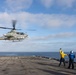A U.S. Marines AH-1Z Viper attack helicopter is hovering just above the flight deck of a naval vessel, likely an amphibious assault ship, during flight operations at sea. Two deck crew members — one in a yellow jersey and one in a blue jersey — are directing the aircraft, with the yellow-jersey handler raising one arm in a signaling gesture. The scene takes place under partly cloudy skies over open ocean.