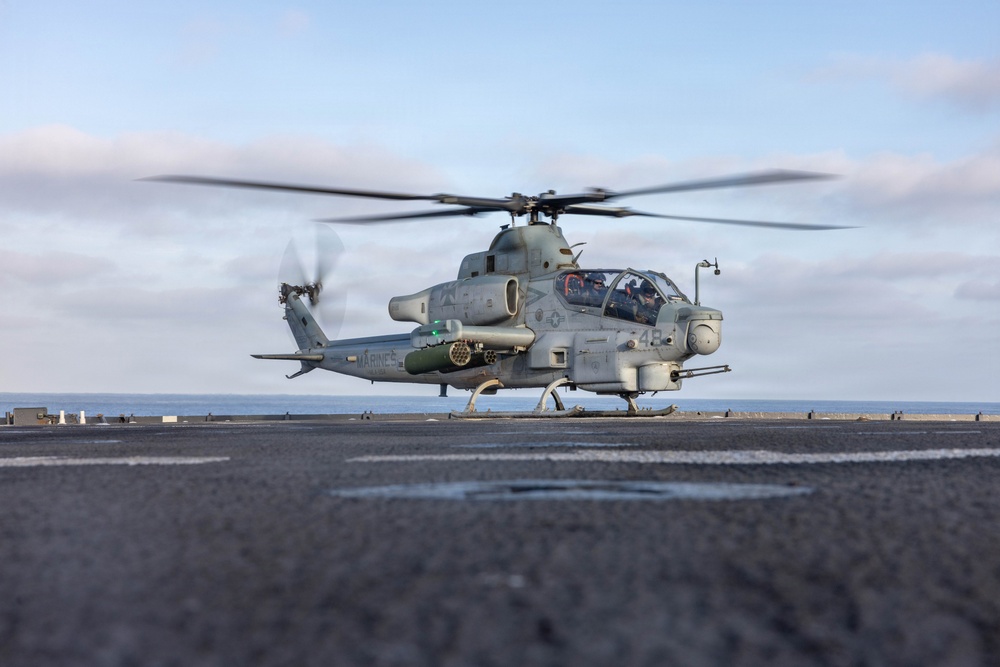 A U.S. Marines AH-1Z Viper attack helicopter is preparing to lift off or has just landed on the flight deck of a naval vessel at sea. The aircraft is armed with weapons systems including what appears to be a Hydra rocket pod and an AIM-9 Sidewinder missile. The rotor blades are in motion, indicating the engines are running.