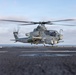 A U.S. Marines AH-1Z Viper attack helicopter is preparing to lift off or has just landed on the flight deck of a naval vessel at sea. The aircraft is armed with weapons systems including what appears to be a Hydra rocket pod and an AIM-9 Sidewinder missile. The rotor blades are in motion, indicating the engines are running.